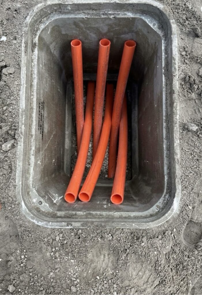 Orange HDPE ducts standing upright in an open handhole over a gravel bed, duct ends open.