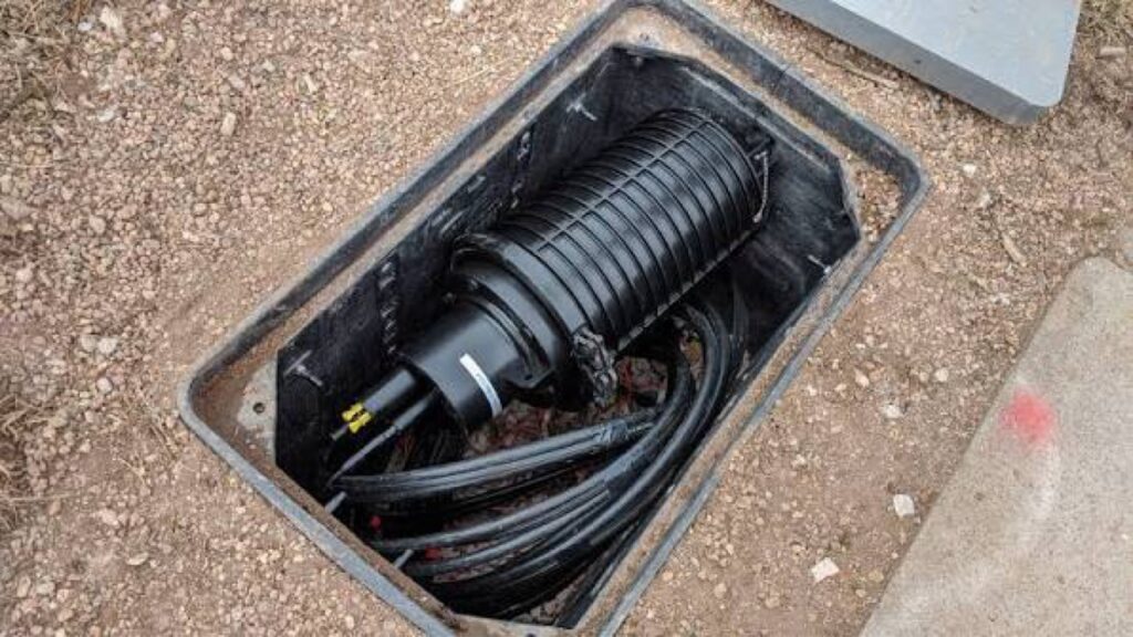 Dome splice closure lying loose in the bottom of a handhole on top of coiled cable.