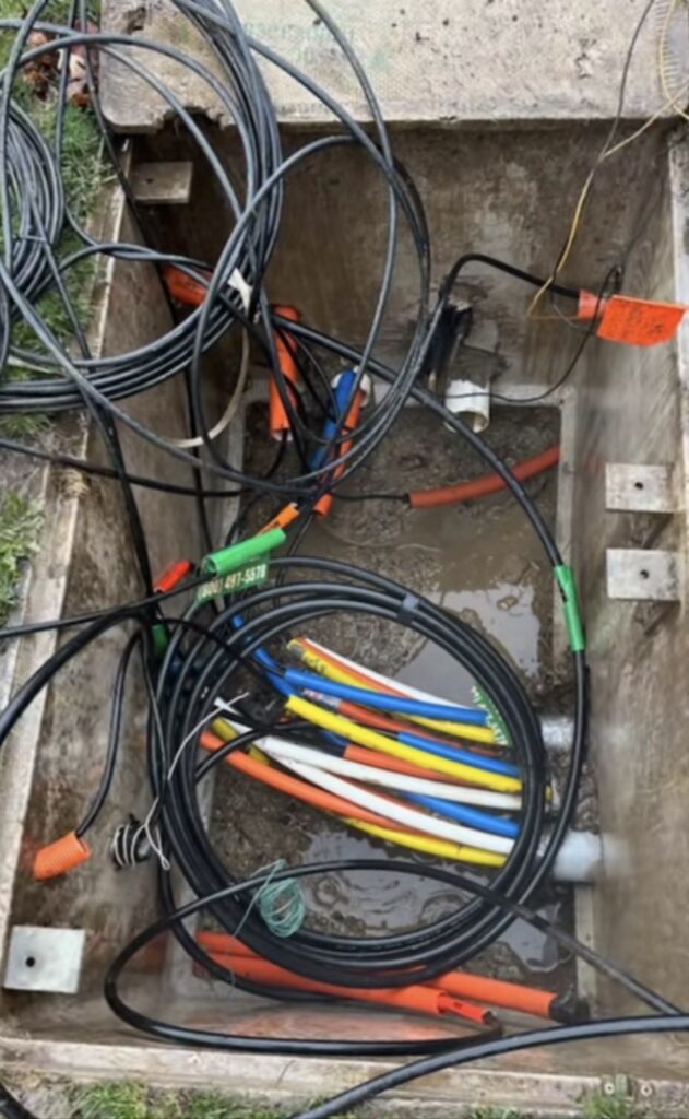 Handhole with standing water in the bottom and cut microducts lying uncapped in the water under a coil of cable.