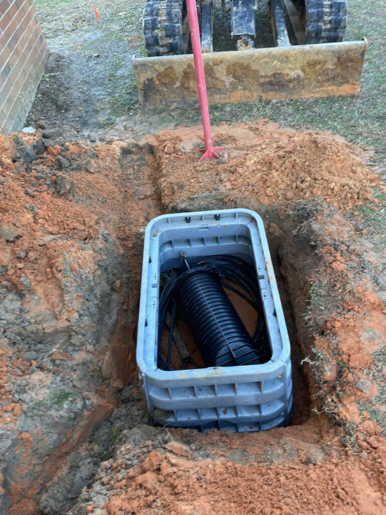 Handhole set in a red dirt excavation with a splice closure and coiled cable inside, mini excavator blade at the top of the frame.